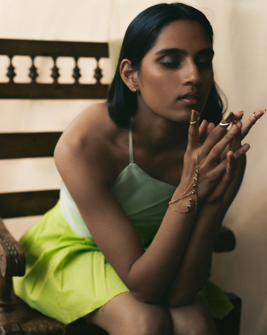 Model sitting on a wood chair in a green top and skirt looking directly at the camera with smokey eye makeup
